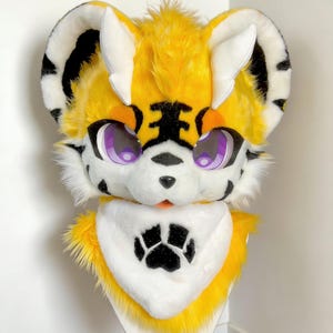 May include: A vibrant, plush tiger fursuit head with yellow, white, and black fur. The head features large, purple eyes, a black nose, and a white bib with a black paw print. The ears are white with black stripes.
