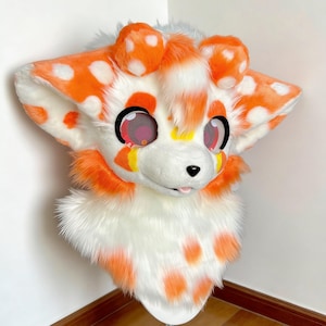 May include: A furry, anthropomorphic head with a white and orange spotted design. The head has large, expressive eyes with pink irises, a small black nose, and a protruding tongue. The ears and cheeks are adorned with orange fur.