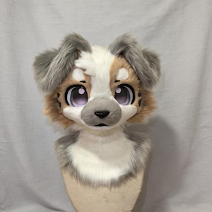 Peut inclure: Un masque de tête de chien en peluche, avec des yeux expressifs et de grandes dimensions. Le pelage est un mélange de blanc, de gris et de beige, avec des détails tels qu'un nez noir et des oreilles touffues. Monté sur un support neutre.