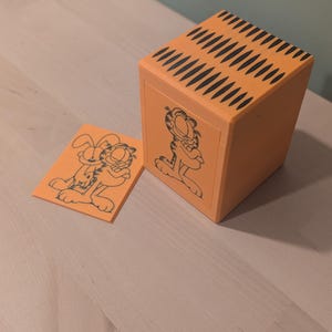May include: An orange rectangular box with black striped patterns on the top surface and a black line drawing of the cartoon character Garfield on the front side. Beside the box lies a small orange rectangular card featuring a black line drawing.