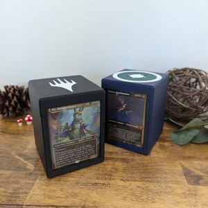 May include: Two black and blue card storage boxes with white and green accents. The box on the left has a white symbol on the top and a card with a rabbit creature on the front. The box on the right has a green symbol on the top and a card with a deer creature on the front. Both boxes have text on the front.