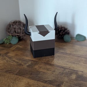 May include: A multi-layered box with a white top, brown middle, and black base. The top features a brown arrow design and is adorned with two black, curved horns, resembling a bison. The box is on a wooden surface.