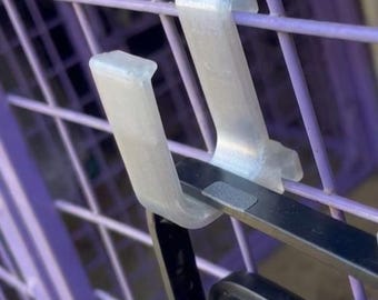 May include: Close-up of two clear plastic clips attached to a purple metal shopping trolley. The clips are designed to hold a black object, possibly a shopping bag. The trolley's metal bars are visible.