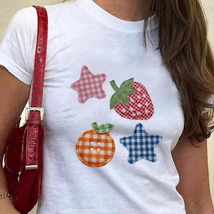 Può includere: T-shirt bianco con applicazioni di frutta e stelle a quadri vichy colorati. La fragola è rossa e bianca, l'arancia è arancione e bianca e le stelle sono rosa e blu. È visibile una borsa rossa con fibbia argentata.