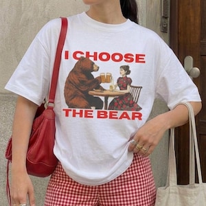 May include: White t-shirt with the text "I CHOOSE THE BEAR" in red. The graphic depicts a bear and a woman at a table with beer. The bear is holding a beer mug. The woman is wearing a floral dress.