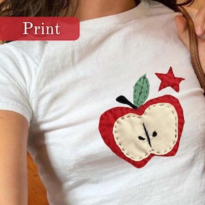 Puede incluir: Camiseta blanca con un aplique de manzana y estrella rojas. El diseño de la manzana presenta un centro color crema, una hoja verde y un tallo negro. La palabra "Print" se muestra en un rectángulo rojo.