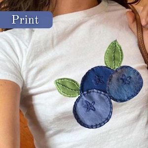 Könnte beinhalten: Weißes T-Shirt mit einem Blaubeer-Design aus Filz. Das Design zeigt drei blaue Blaubeeren mit gestickten Sterndetails und grünen Blättern. Das Wort "Print" befindet sich in einem blauen Rechteck oben links.