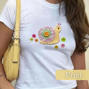 May include: White t-shirt with a snail applique. The snail has a yellow body and a pink and green shell, decorated with colourful buttons. The word "Print" is displayed in a yellow rectangle at the bottom of the image.
