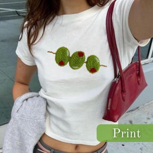 Puede incluir: Camiseta blanca con un diseño bordado de tres aceitunas verdes con centros rojos en un pincho de madera. La camiseta es de manga corta y tiene cuello redondo. Se ve un bolso rojo. La palabra "Print" está en un rectángulo verde.