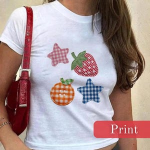 Puede incluir: Camiseta blanca con aplicaciones de frutas y estrellas de cuadros de colores. El diseño incluye una estrella rosa, una fresa roja y blanca, una naranja naranja y blanca y una estrella azul. La palabra "Print" está en un rectángulo rojo.