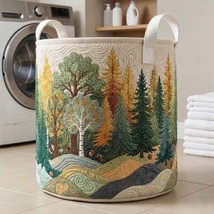 May include: A round laundry hamper with a quilted forest scene. The design features trees in shades of green, yellow, and brown, with a cream-colored background. The hamper has white handles and is placed near a washing machine and folded towels.