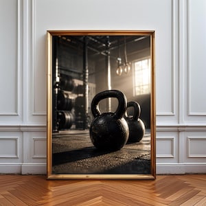 May include: A framed wall art print featuring two black kettlebells resting on a gym floor. The background shows a blurred fitness facility with weight racks and hanging gymnastic rings illuminated by natural light streaming through a window.