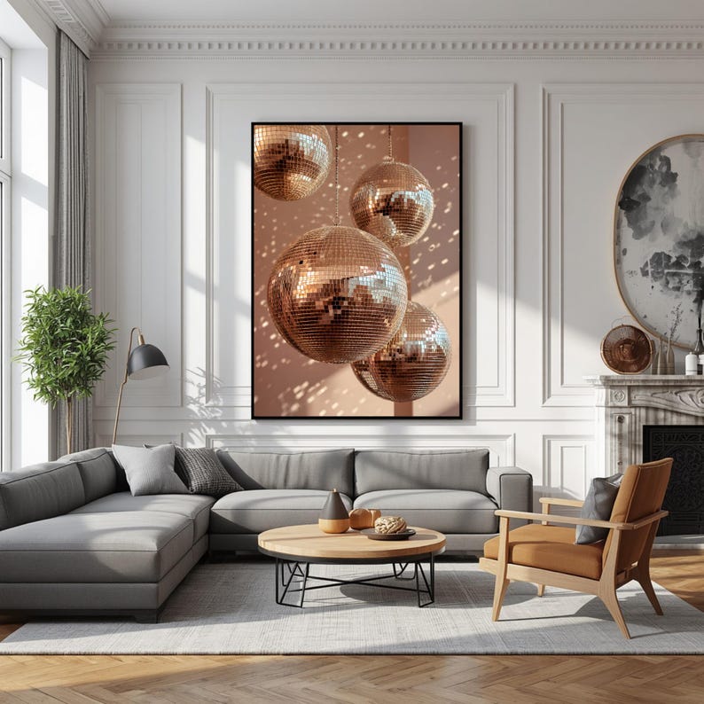 K&ouml;nnte beinhalten: Ein rechteckiger Wandkunstdruck mit drei reflektierenden ros&eacute;goldenen Discokugeln, die vor einem sanften pfirsichfarbenen Hintergrund h&auml;ngen und ein Muster aus Lichtpunkten erzeugen. Das Kunstwerk ist schwarz gerahmt an einer wei&szlig;en Wand.