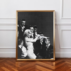 May include: A framed black and white photograph showing a group of people, including a woman in a light-coloured dress and long gloves, surrounded by men in tuxedos. The photograph is in a gold frame and is displayed on a wooden floor.