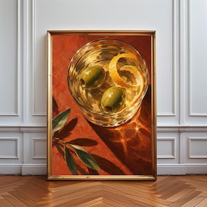 Op de afbeelding: Een ingelijste kunstdruk met een bovenaanzicht van een helder glas gevuld met een gouden vloeistof, twee groene olijven en een gekrulde sinaasappelschil. De achtergrond is warm terracotta oranje, met een kleine groene olijftak onderaan.