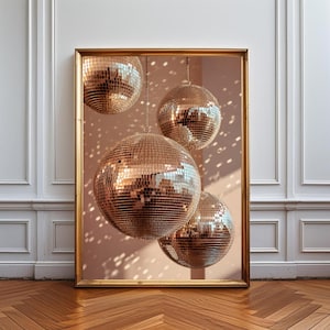 K&ouml;nnte beinhalten: Ein gerahmter Kunstdruck mit vier kugelf&ouml;rmigen Discokugeln mit reflektierenden goldenen Spiegeloberfl&auml;chen. Die Discokugeln h&auml;ngen in unterschiedlichen H&ouml;hen vor einem sanften pfirsichfarbenen Hintergrund und erzeugen verstreute Lichtreflexe.