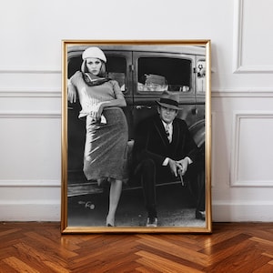 Könnte beinhalten: Gerahmter Schwarz-Weiß-Druck mit einer Vintage-Fotografie eines Mannes und einer Frau in Kleidung der 1930er Jahre. Der Mann, in Anzug und Hut, sitzt, während sich die Frau an ein Auto lehnt. Der Druck ist in einem goldenen Rahmen.