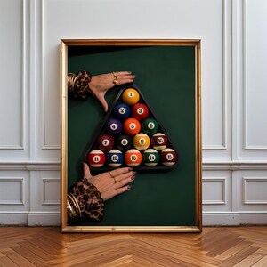 May include: A framed wall art print featuring a triangular rack of fifteen numbered billiard balls arranged on a dark green surface. Two hands with gold rings and leopard print sleeves are positioned at the top and bottom of the black plastic rack.
