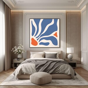 May include: A square wall art piece featuring an abstract design with organic, flowing blue shapes on a cream background, accented by two solid orange shapes, all enclosed within a thin black frame, displayed above a bed in a bedroom.