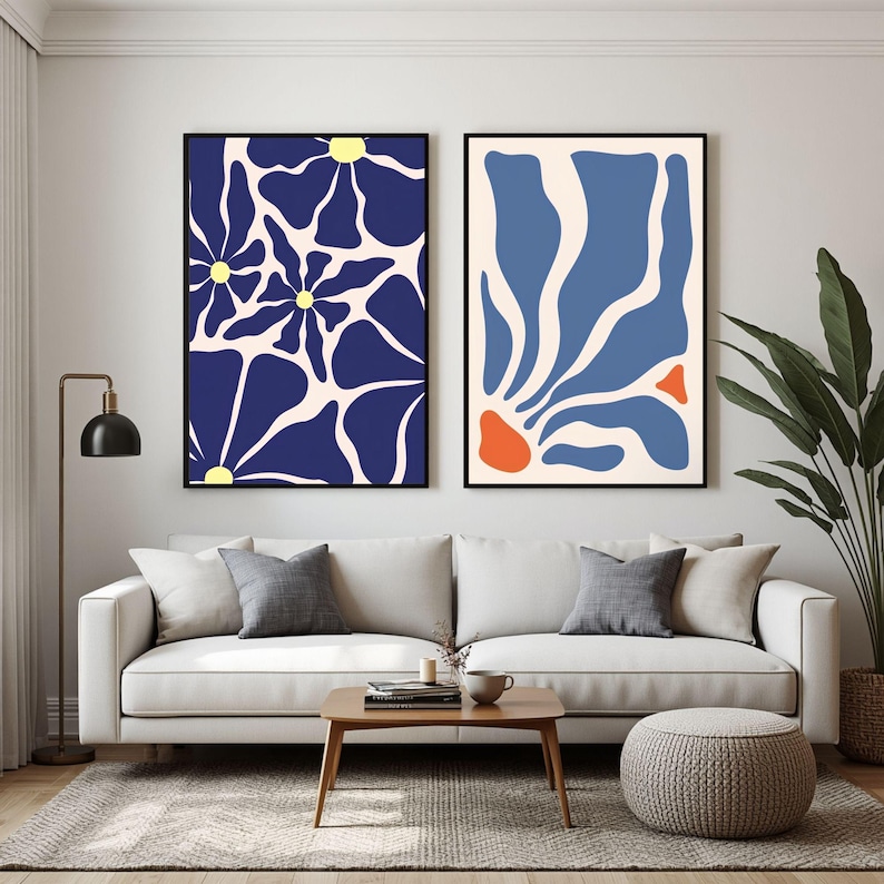 May include: Two framed rectangular wall art prints hanging side by side. The left print features dark blue abstract flower shapes with yellow centres on a cream background. The right print displays blue and orange organic shapes on a cream background.