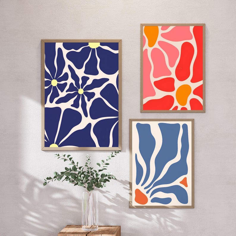 May include: Three rectangular framed wall art prints. The left print features dark blue flower shapes with yellow centres on a cream background. The top right print shows red, pink, and orange organic shapes. The bottom right print displays blue and orange shapes.