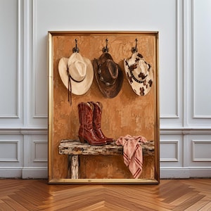 Op de afbeelding: Een ingelijst schilderij met drie cowboyhoeden aan wandhaken boven een paar bruine leren cowboylaarzen op een rustieke houten bank, met een gevouwen lichtgekleurde stof gedrapeerd over de rand van het bankoppervlak.