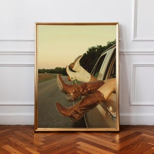 Può includere: Stampa incorniciata con due paia di stivali da cowboy che sporgono dal finestrino di un'auto. Un paio è marrone, l'altro bianco. Lo sfondo presenta una strada e un tramonto.