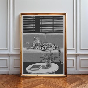 Peut inclure: Une impression d'art encadr&eacute;e en noir et blanc repr&eacute;sentant un l&eacute;opard allong&eacute; sur un canap&eacute;. Un verre &agrave; martini est pos&eacute; sur le coussin &agrave; c&ocirc;t&eacute; du l&eacute;opard. Au premier plan, une table ronde porte un petit vase avec des feuilles et deux livres.