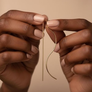 May include: A pair of gold-colored earrings. One earring is a straight, thin bar, and the other is a curved, flowing design. The earrings are held up by a person's hands, showcasing the jewelry's delicate design and color.