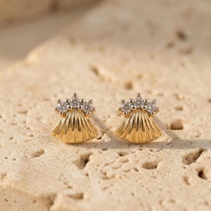 May include: A pair of gold stud earrings. Each earring has a fan-shaped design with a textured surface and a cluster of small, clear gemstones at the top. The earrings are presented on a light brown, textured surface.