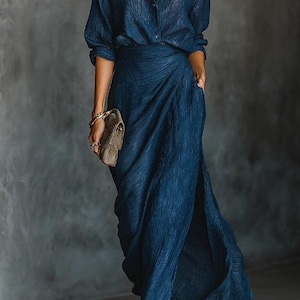 May include: A dark blue, long-sleeved shirt and matching skirt. The skirt has a high slit and a wrap design. The outfit is paired with a small, brown clutch. The fabric has a textured appearance.