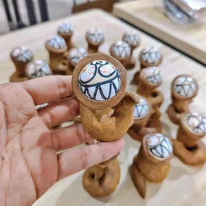 May include: A collection of whimsical, brown ceramic figurines. Each has a round head with a cartoonish face featuring large eyes and a wide, toothy grin. The bodies are uniquely shaped, with a curved, arm-like appendage.