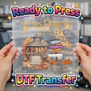 May include: A clear DTF transfer sheet featuring a colorful graphic with the text Ready to Press and DTF Transfer. The design includes a gazebo, a bench, pumpkins, and a signpost with the text Stars Hollow, Est 1779, Dragonfly Inn, Luke's Diner, Antiques, and Where You Lead I Will Follow.