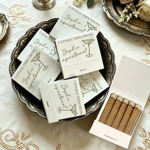 May include: A collection of white matchboxes featuring the text I STOLE THIS FROM Sophia's apartment and a martini glass illustration in dark green ink. One open matchbox displays a row of wooden matches with light brown tips inside the box.