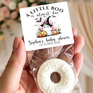 May include: A square white card topper with the text A LITTLE BOO is almost due, Sophia's baby shower, and OCTOBER 2026. The design features a ghost in a witch hat, two orange pumpkins, and bats. A clear plastic bag contains a white ring-shaped candy.