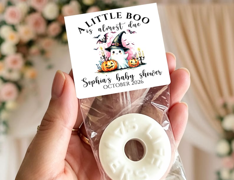 May include: A square white card topper with the text A LITTLE BOO is almost due, Sophia's baby shower, and OCTOBER 2026. The design features a ghost in a witch hat, two orange pumpkins, and bats. A clear plastic bag contains a white ring-shaped candy.