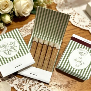 May include: Three matchbooks with a green and white vertical stripe pattern. One open matchbook reveals wooden matches with white tips. The closed matchbooks feature circular labels with line art of cupids, hearts, and the text Sydney & Kyle May 16, 2026.