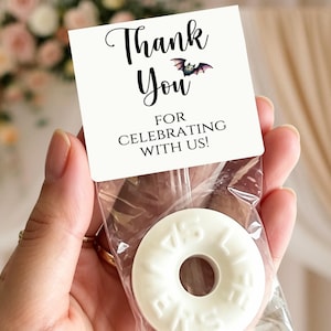 May include: A clear plastic favor bag containing a white ring-shaped candy with embossed text. The bag is sealed with a white cardstock topper featuring the black printed text Thank You For Celebrating With Us and a small illustration of a bat.