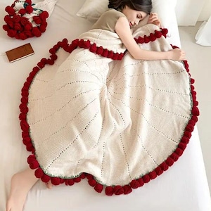 Puede incluir: Una manta de punto color crema con un ribete de pompón rojo. La manta tiene un diseño circular con líneas radiales. Un ramo de rosas rojas y un pequeño cuaderno también están en la cama.