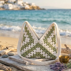 Puede incluir: Bolso tote de crochet hecho a mano con un patrón geométrico en crema y verde oliva. El bolso tiene largas correas color crema y se muestra sobre una superficie de madera cerca de la playa. El fondo presenta el océano y una ciudad costera.