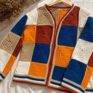 K&ouml;nnte beinhalten: Eine lang&auml;rmelige Strickjacke, die aus einzelnen quadratischen H&auml;kelst&uuml;cken in einer lebendigen Farbpalette aus K&ouml;nigsblau, leuchtendem Orange, dunklem Rostrot, Beige und Wei&szlig; gefertigt ist. Das Kleidungsst&uuml;ck hat eine offene Vorderseite.