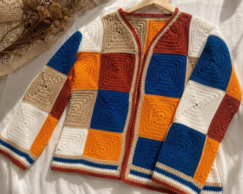 K&ouml;nnte beinhalten: Eine lang&auml;rmelige Strickjacke, die aus einzelnen quadratischen H&auml;kelst&uuml;cken in einer lebendigen Farbpalette aus K&ouml;nigsblau, leuchtendem Orange, dunklem Rostrot, Beige und Wei&szlig; gefertigt ist. Das Kleidungsst&uuml;ck hat eine offene Vorderseite.