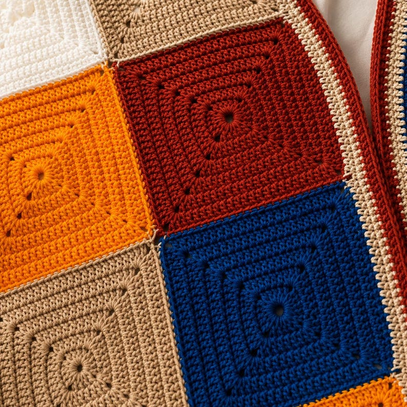 K&ouml;nnte beinhalten: Ein H&auml;kelkleidungsst&uuml;ck, das aus mehreren quadratischen Motiven in Unifarben wie Dunkelrot, leuchtendem Orange, K&ouml;nigsblau, Beige und Wei&szlig; besteht. Die Quadrate sind mit einem cremefarbenen Rand verbunden und zeigen ein detailliertes Garnmuster.
