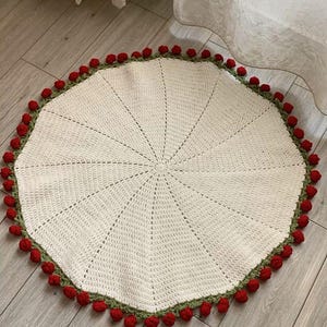 May include: A round, off-white crochet rug with a green border and red pom-poms. The rug has a radial design, with lines radiating from the center. This handmade rug is a decorative home accent.