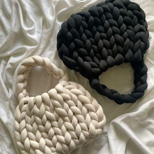 May include: Two handmade, chunky knit bags. One is off-white, the other is black. Both have a rounded shape with braided handles. The bags are made of thick, soft yarn and sit on a white, wrinkled fabric background.