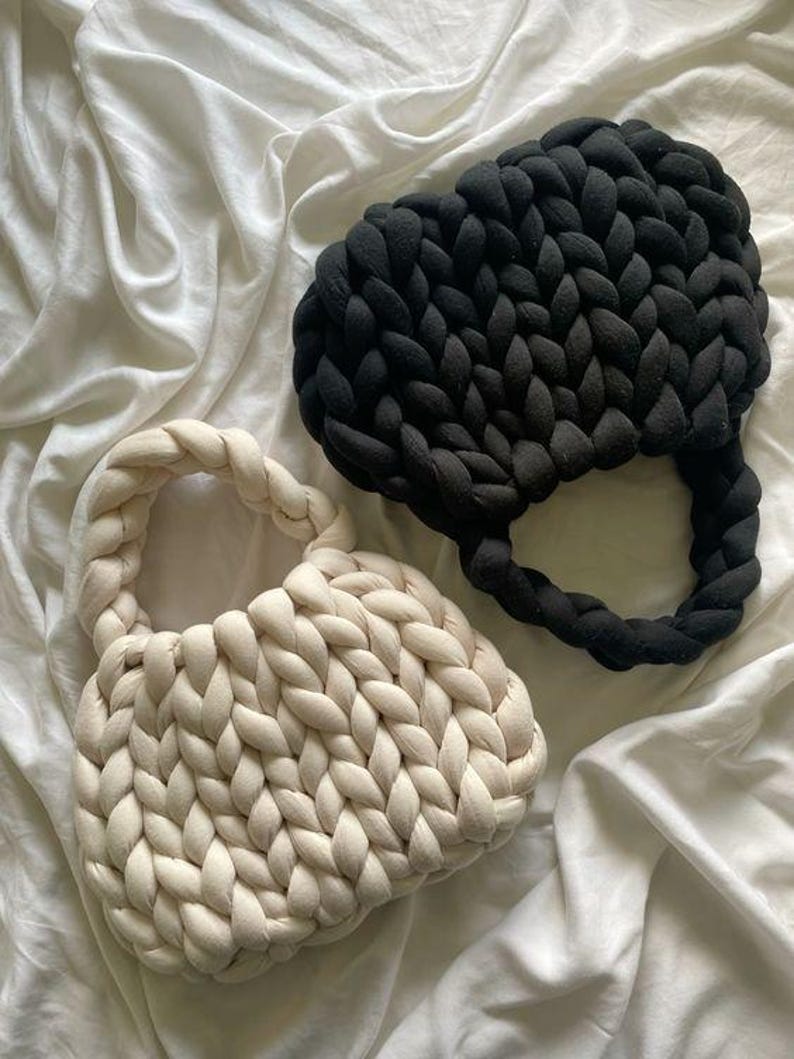 May include: Two handmade, chunky knit bags. One is off-white, the other is black. Both have a rounded shape with braided handles. The bags are made of thick, soft yarn and sit on a white, wrinkled fabric background.