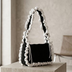 以下が含まれることがあります： 黒いかぎ針編みのハンドバッグ。長方形の本体の縁とショルダーストラップに沿って白いフリルのトリムが施されています。バッグの両サイドには白い引き紐が付いており、無地の黒いベースに対して質感のあるコントラストを生み出しています。
