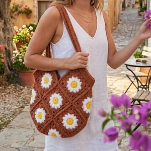 Könnte beinhalten: Eine braune Häkeltasche mit weißen und gelben Gänseblümchenmustern. Die Tasche hat einen Schulterriemen. Der Hintergrund umfasst Blumen und einen Steinweg.