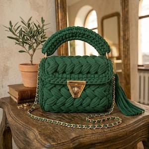 Op de afbeelding: Een handgemaakte, smaragdgroene handtas met een gevlochten ontwerp. De tas heeft een goudkleurige sluiting, een kettingriem en een kwast. De handgreep is ook gevlochten. De tas staat op een houten tafel.