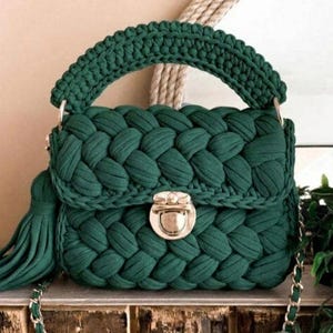 May include: A dark green crochet handbag with a distinctive braided pattern. It has a top handle, a flap secured by a gold-coloured clasp, and a long tassel. A silver chain strap is also visible.